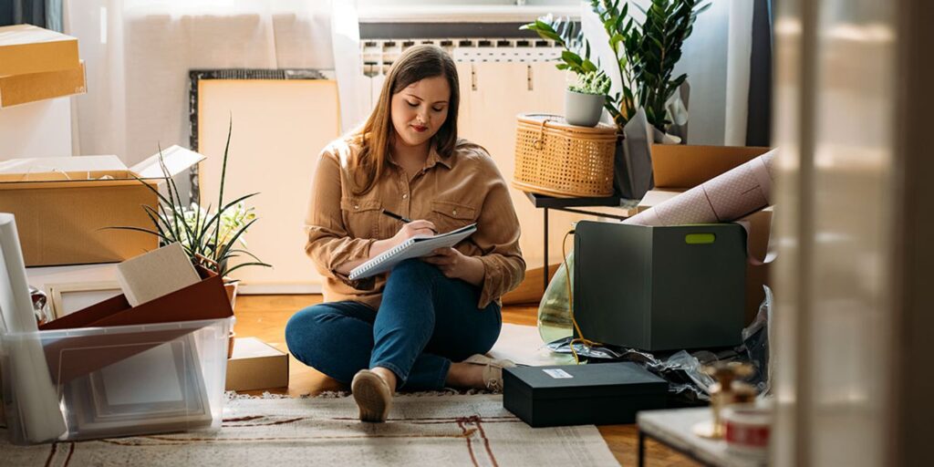37fc097c8d87-19-moving-house-checklist Woman sitting among moving boxes, writing in a notebook, surrounded by plants and home decor. | Sky Rye Design Woman sitting among moving boxes, writing in a notebook, surrounded by plants and home decor.