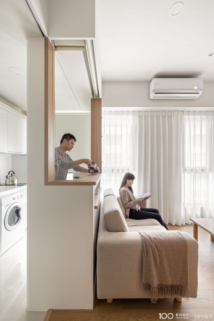 Modern apartment interior with a man in the kitchen and a woman reading on the sofa under natural light.