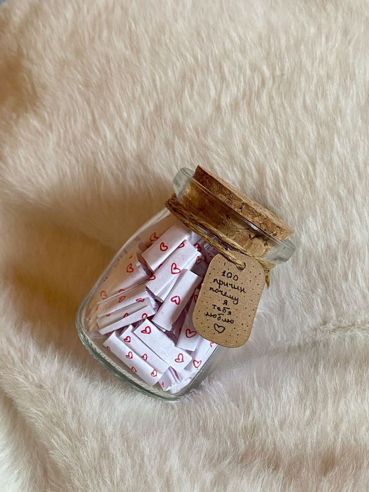 Glass jar filled with love notes and a cork lid, tagged with romantic message on a soft, fluffy surface.