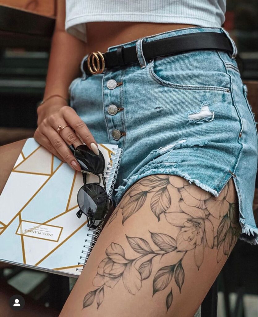 Close-up of woman's tattooed leg in ripped denim shorts, holding a planner and sunglasses.