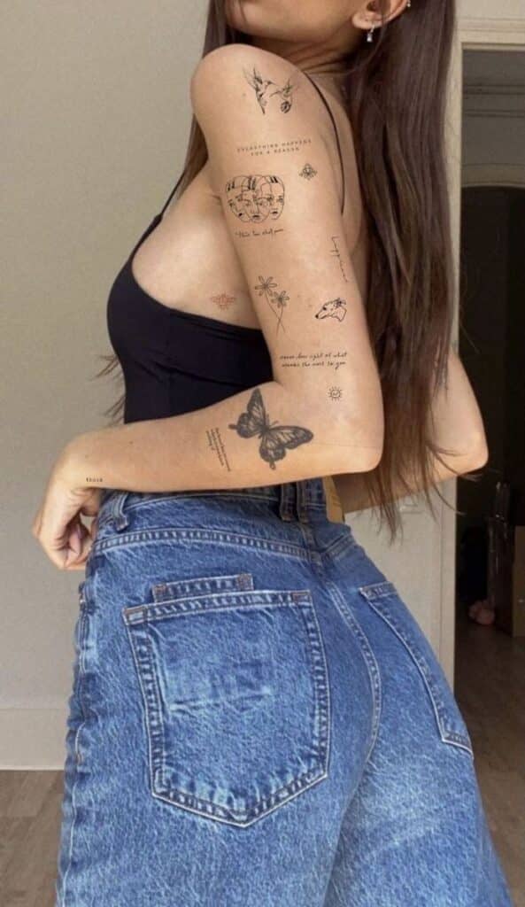 Woman in a black top and jeans showing arm tattoos featuring various designs and text, standing in a casual pose.