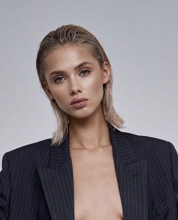 Young person with wet slicked-back hair, wearing a pinstriped blazer, posing against a neutral background.