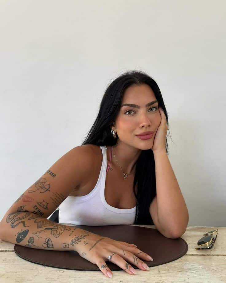 Smiling woman with tattoos sitting at table, resting her chin on hand, wearing white tank top and accessorized jewelry.