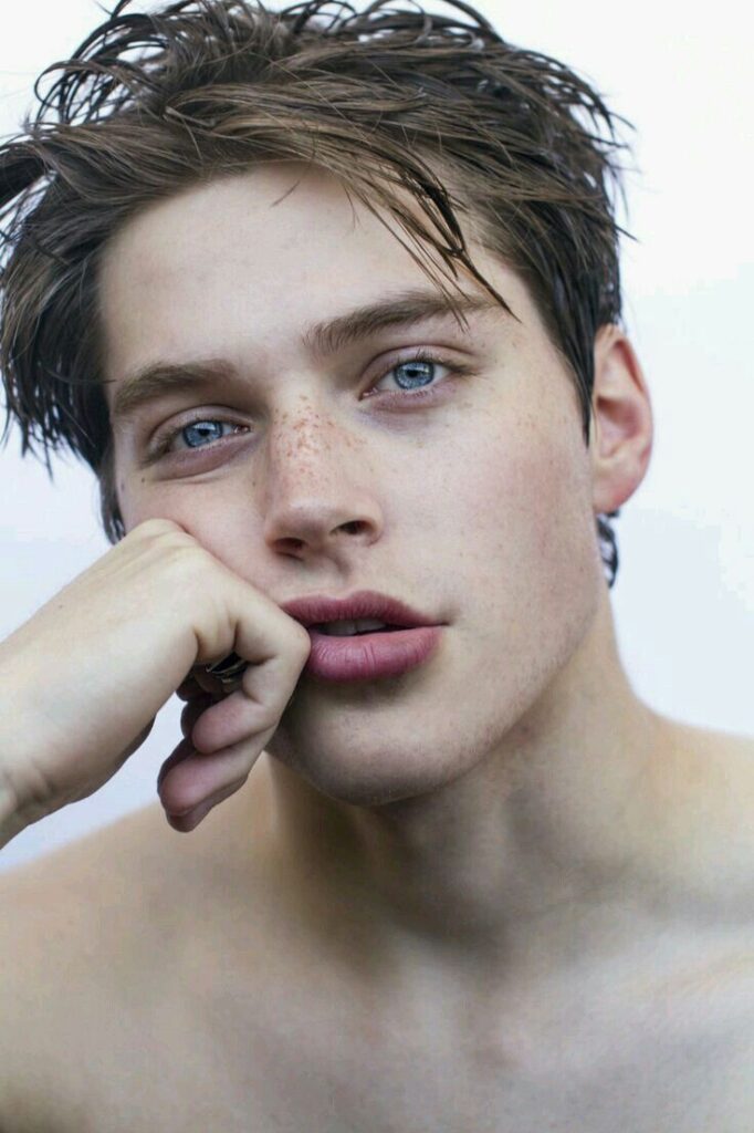 Portrait of a young person with blue eyes and tousled hair, resting their chin on their hand, looking thoughtful.