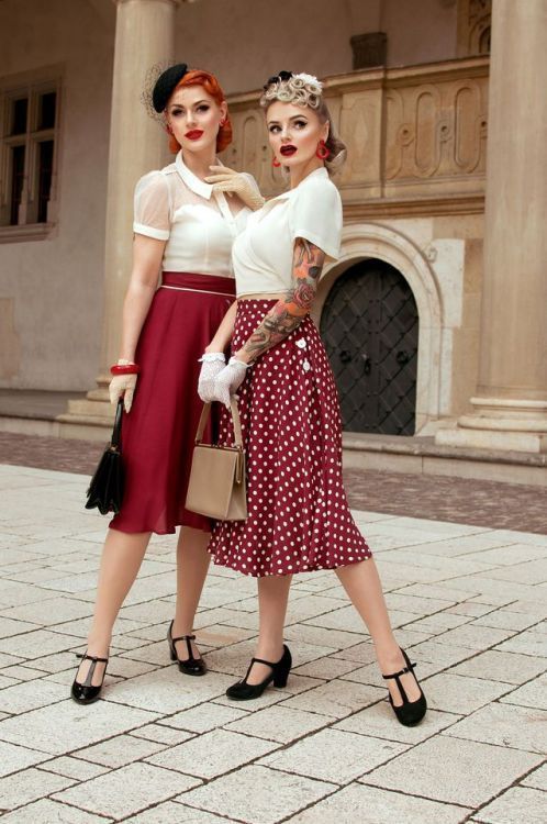 Two women in vintage outfits with red skirts and retro accessories posing stylishly in front of an elegant building.