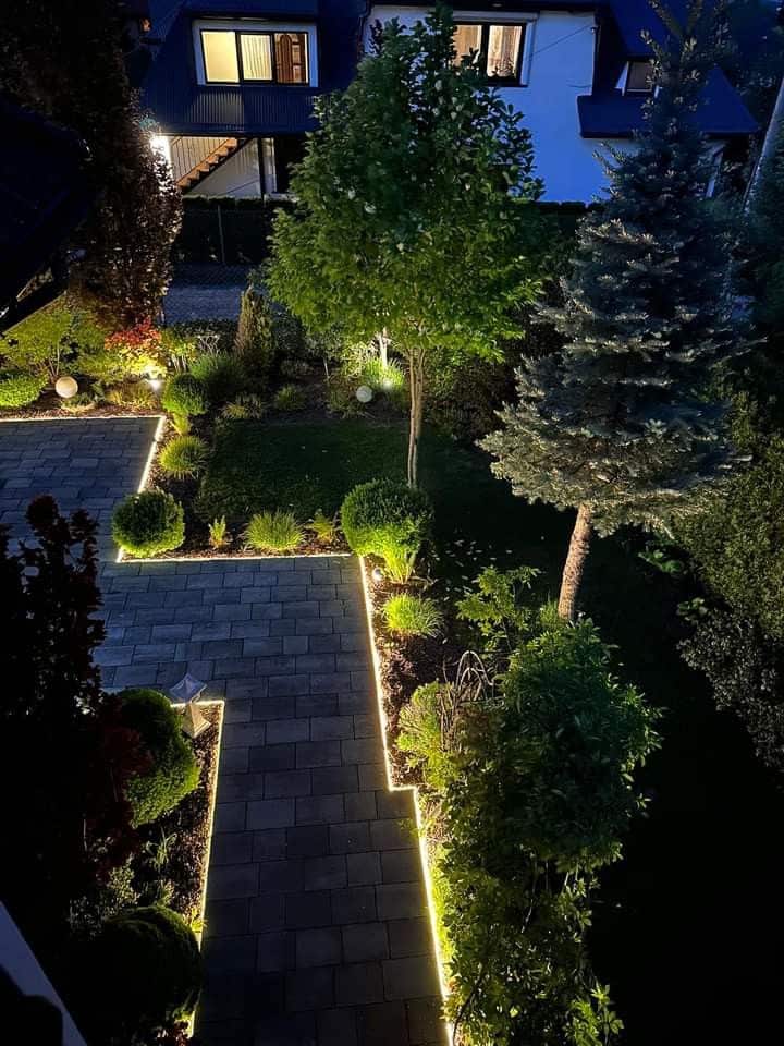 Illuminated garden path with trees and lights leading to a house at night.