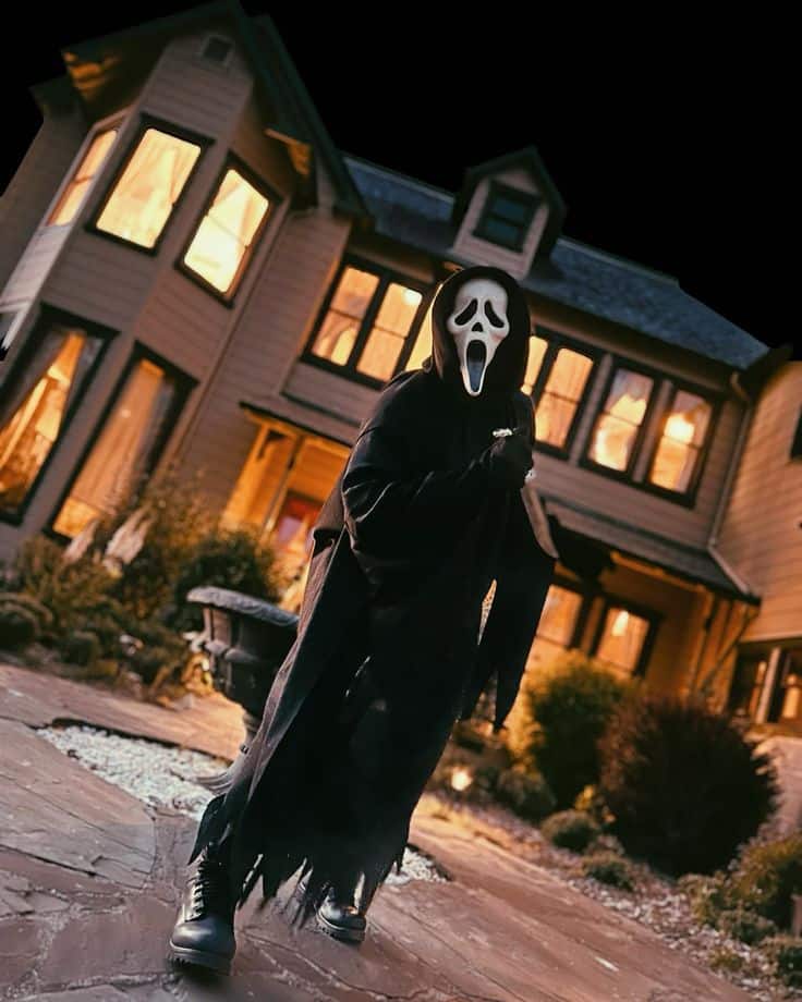 Person in a ghost mask and black robe stands in front of a lit-up house at night, creating a spooky Halloween scene.