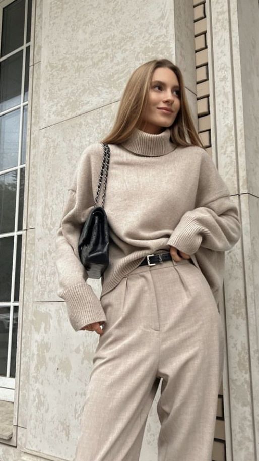 Woman in chic beige outfit with turtleneck and trousers, holding a black bag, standing by a modern building facade.