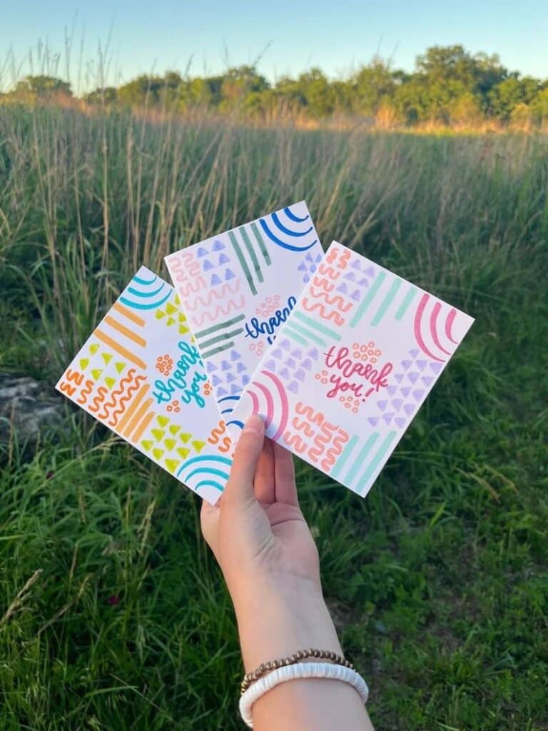 41ce598c32a608fbb3f9a79f38a80787 Hand holding colorful thank you cards with vibrant patterns against a grassy field background. | Sky Rye Design Hand holding colorful thank you cards with vibrant patterns against a grassy field background.