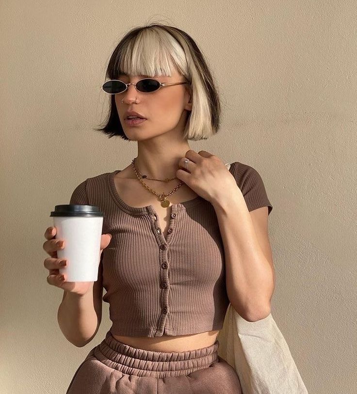 Fashionable woman in cropped shirt holds coffee cup, wearing sunglasses and unique hairstyle, against neutral background.