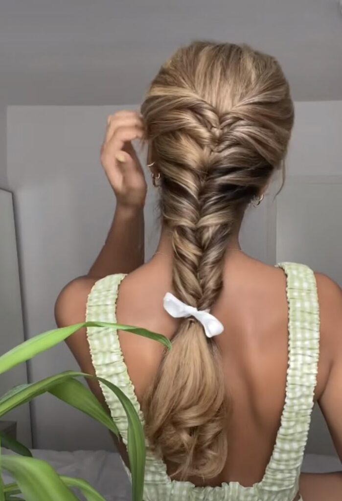 Woman with fishtail braid hairstyle tied with white ribbon, wearing green dress, viewed from the back.