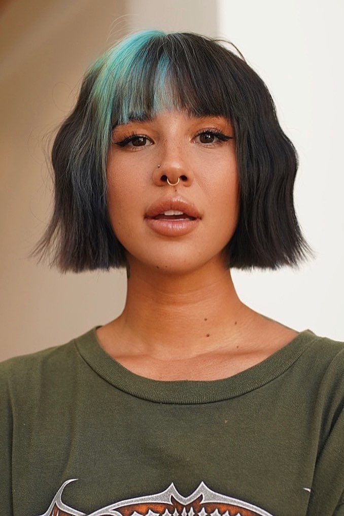 Woman with short black hair, green highlights, wearing a green shirt and septum ring, looking confidently at the camera.