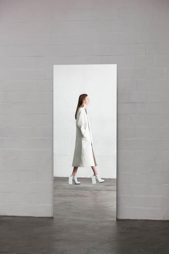 Woman in white coat and boots walking past a mirror against a white brick wall. Stylish modern minimalist fashion.
