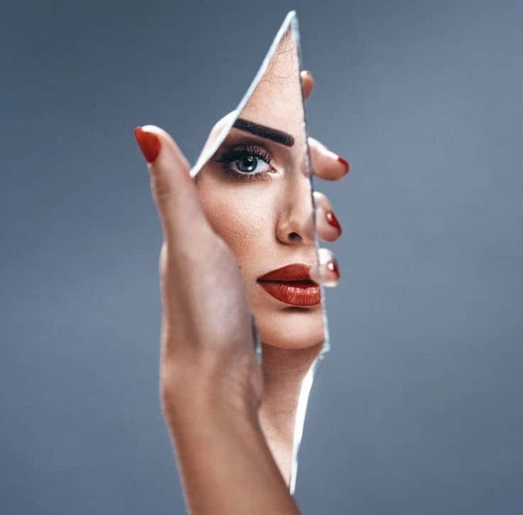 Woman's face reflected in broken mirror piece, red lipstick and nails, blue background.