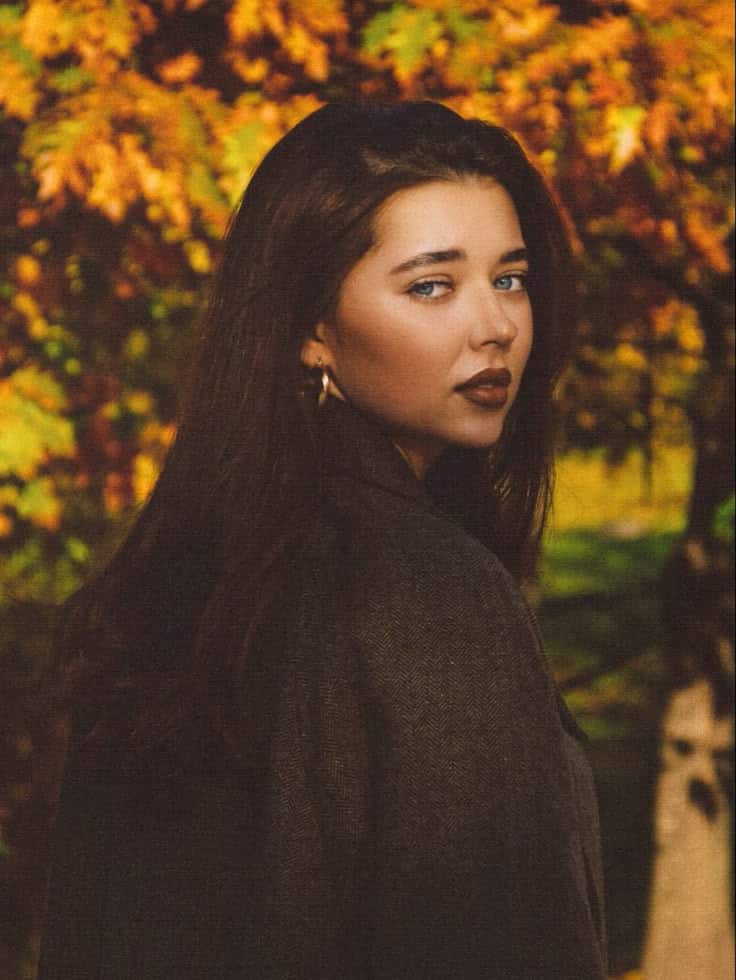 Woman in autumn scenery with vibrant orange leaves, wearing a dark coat, gazes thoughtfully over her shoulder.