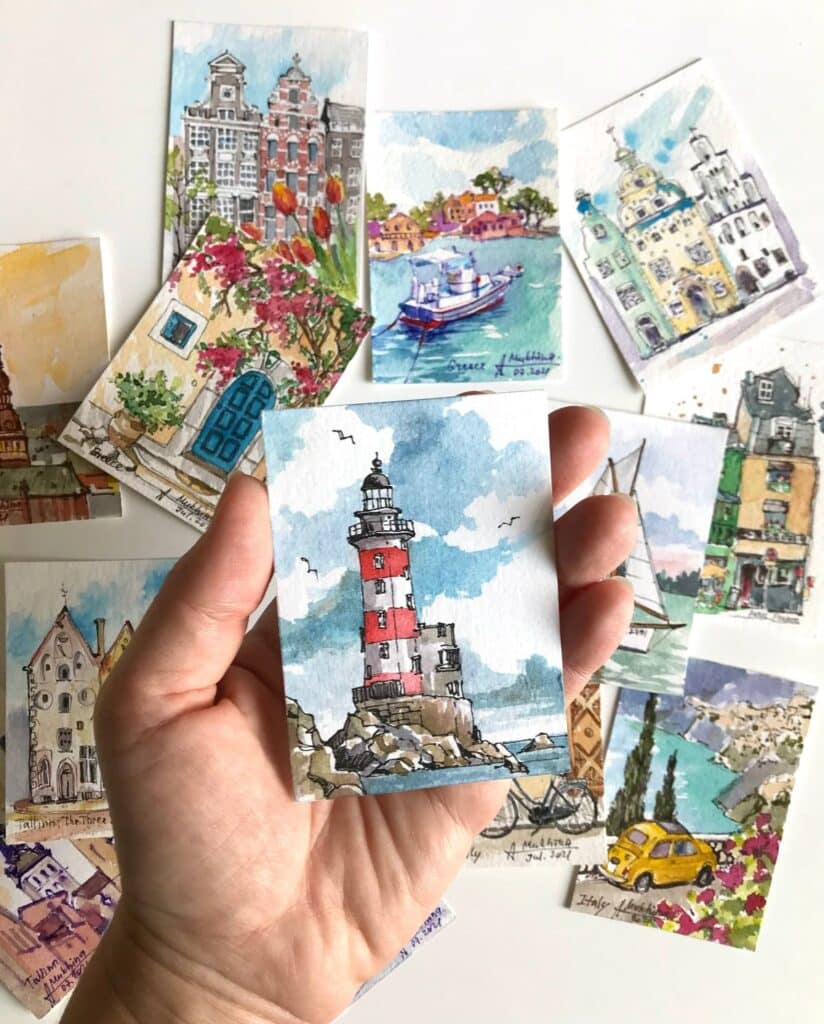 Hand holding watercolor postcards of scenic landscapes and architecture, featuring a lighthouse in the foreground.