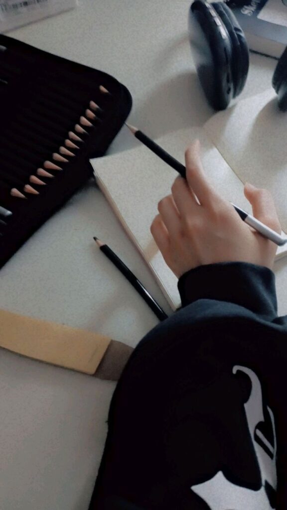 Person sketching in a notebook with pencils and headphones on a desk. Artistic workspace setup.