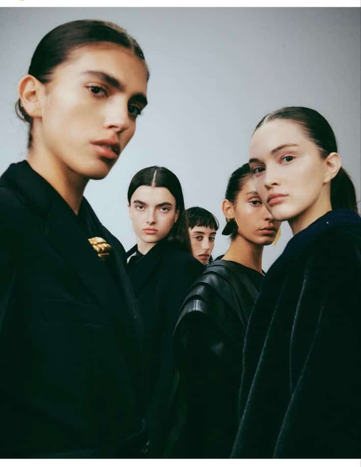 Five models in black fashion attire pose with serious expressions against a neutral background.