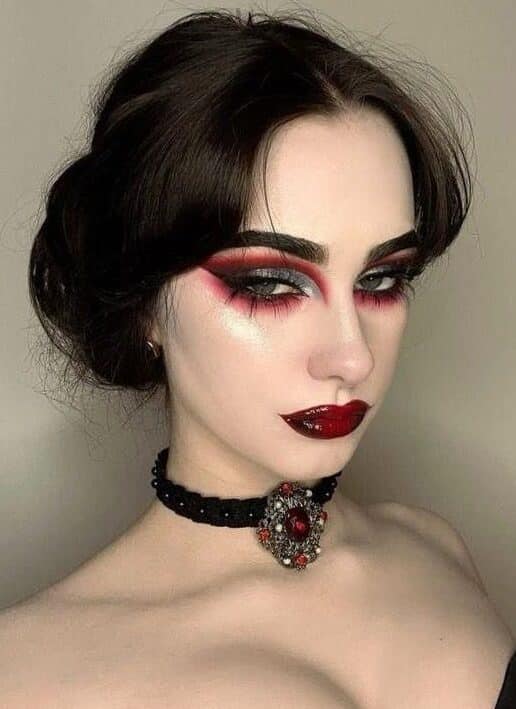 Woman with dramatic red and black makeup, wearing a gem choker and strapless black outfit, posing confidently.