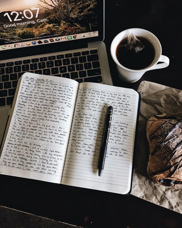 Open laptop, notebook with writing, pen, coffee, and pastry on a table, perfect for a cozy work or study session.