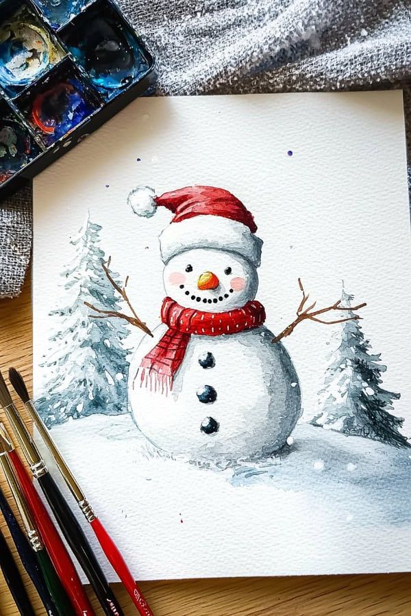 Watercolor painting of a cheerful snowman with red scarf and hat, surrounded by snowy trees and paintbrushes nearby.