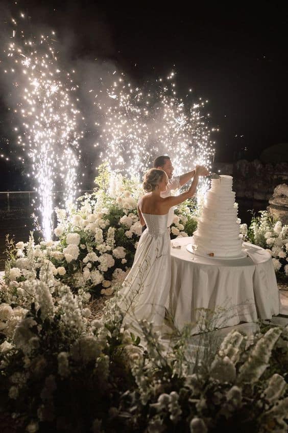 Bride and groom cutting a tiered wedding cake surrounded by fireworks and white flowers in a nighttime garden setting.