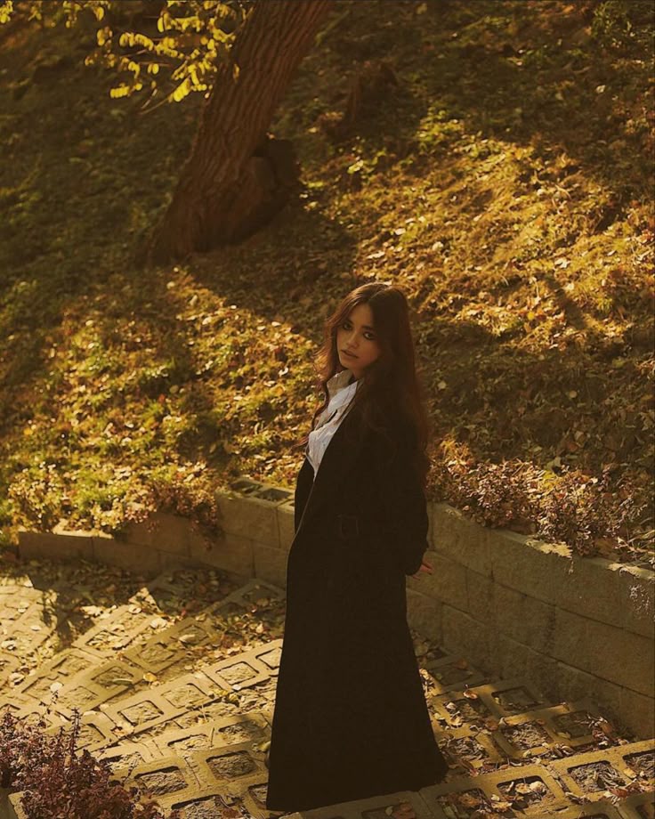Woman in long coat standing on sunlit pathway amidst autumn leaves and trees.