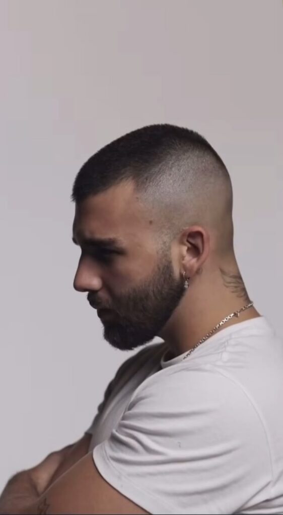 Profile of a man with a buzz cut and beard, wearing earrings and a white shirt, against a neutral background.