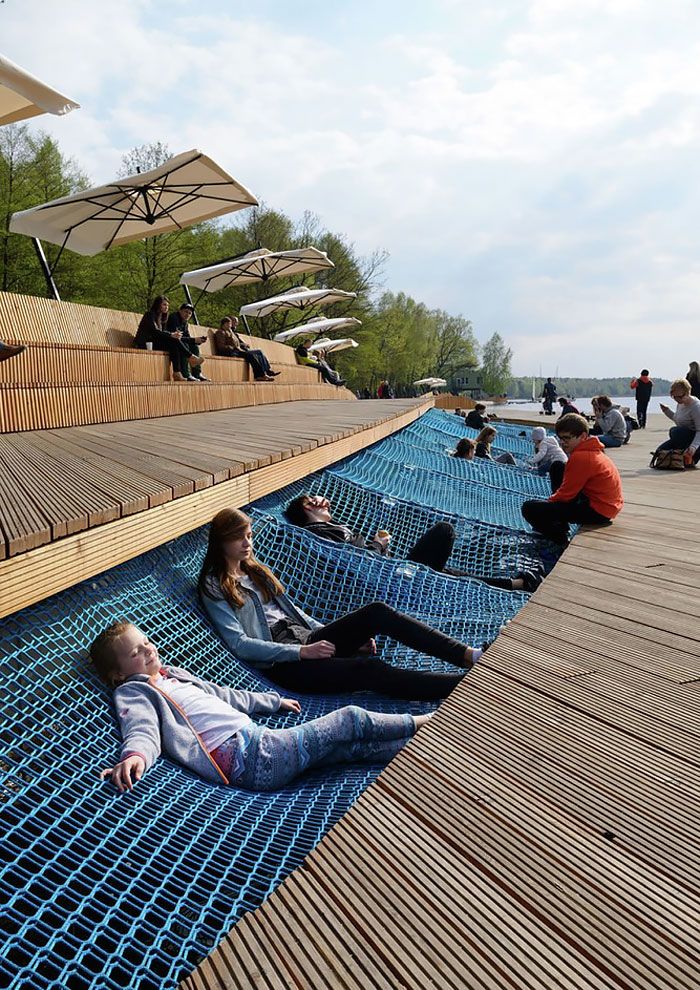 People relax on blue netting by a wooden deck with umbrellas, enjoying a sunny day outdoors near a waterfront.