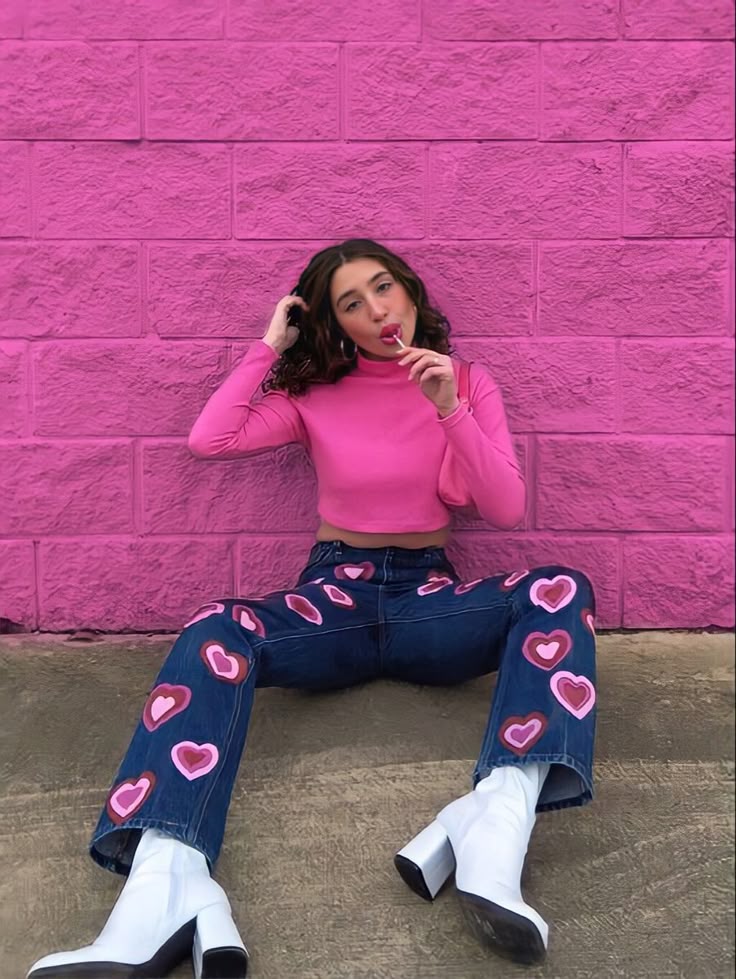 Person in pink top and heart-patterned jeans sits against pink wall, holding a lollipop, wearing white boots.