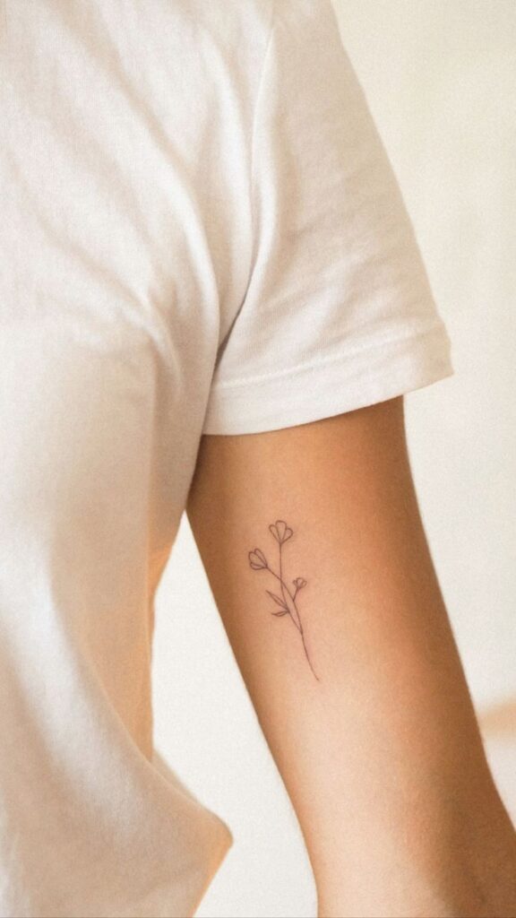 Minimalist flower tattoo on arm in simple black ink, complementing white t-shirt.