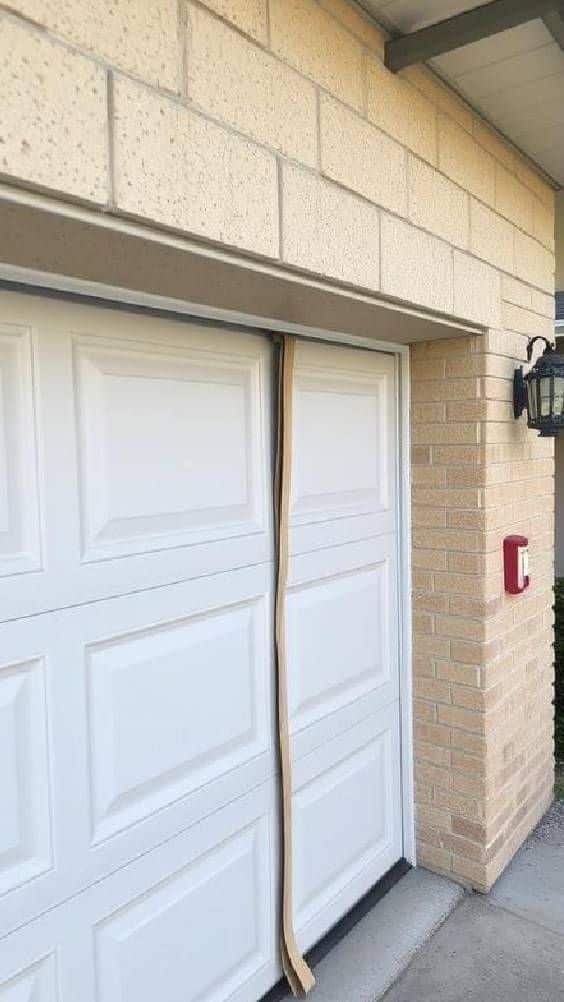 White garage door slightly open with wooden panel sticking out, brick wall and outdoor lantern visible.