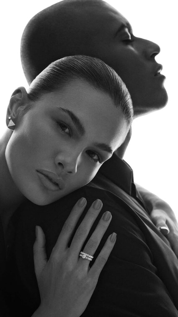 Couple posing elegantly, woman wearing diamond ring and earrings, embracing tenderly in black and white portrait.