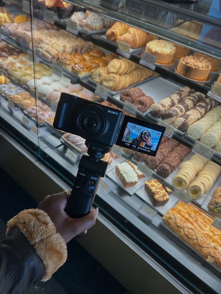 Vlogger filming delicious pastries in a bakery display case with a camera.