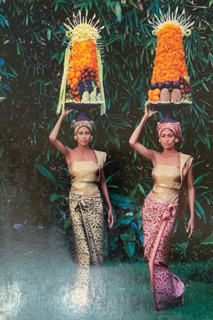 Balinese women in traditional attire carrying colorful offerings on their heads surrounded by lush greenery.