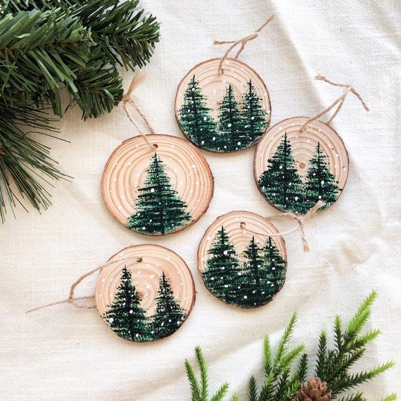 52092e2e-c5ac-41fe-92e9-dc521e448779 Wood slice ornaments with painted evergreen trees and snowflakes on a white cloth background, surrounded by greenery. | Sky Rye Design Wood slice ornaments with painted evergreen trees and snowflakes on a white cloth background, surrounded by greenery.