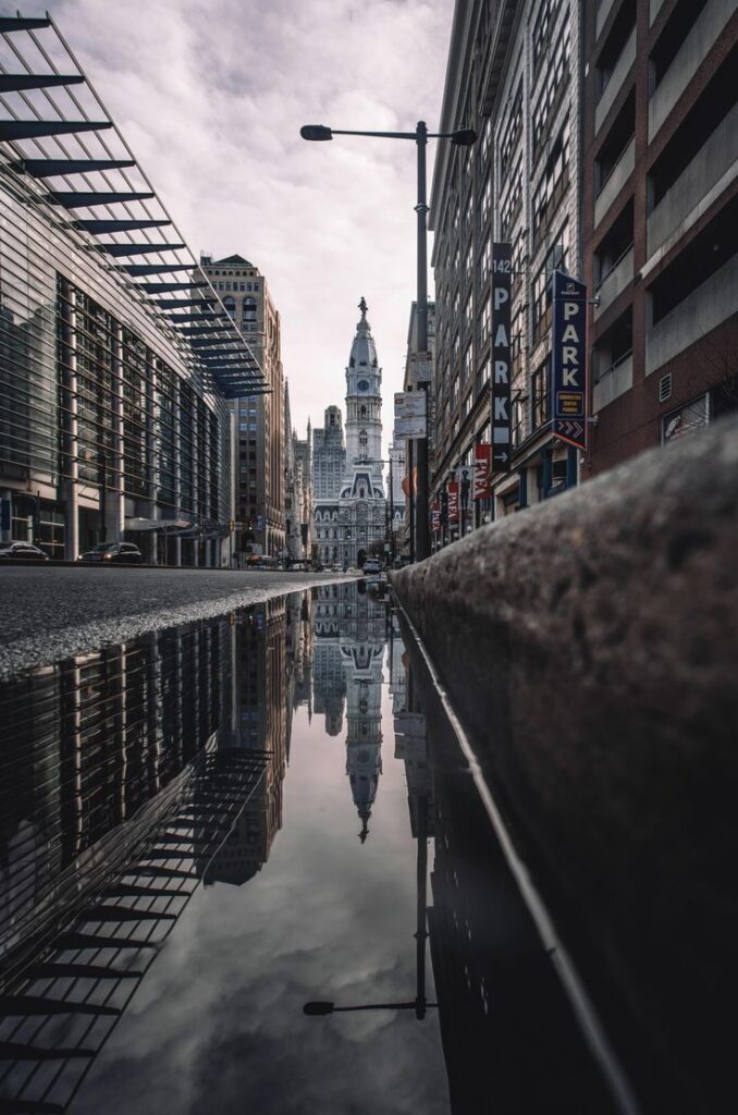 Urban street view with tall historic building reflected in puddle, surrounded by skyscrapers and cloudy sky.