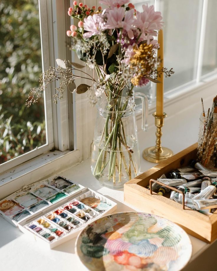 Sunny art studio with flowers, watercolor paints, and brushes on a windowsill, perfect for creativity.