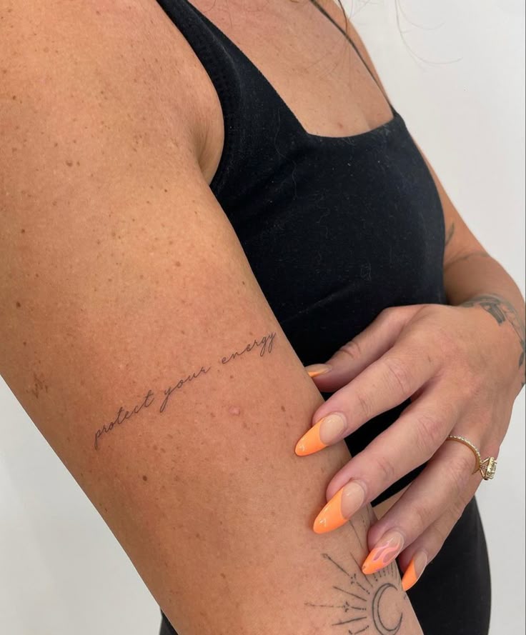 Arm with protect your energy tattoo, person in black top, orange nails, sun tattoo visible.