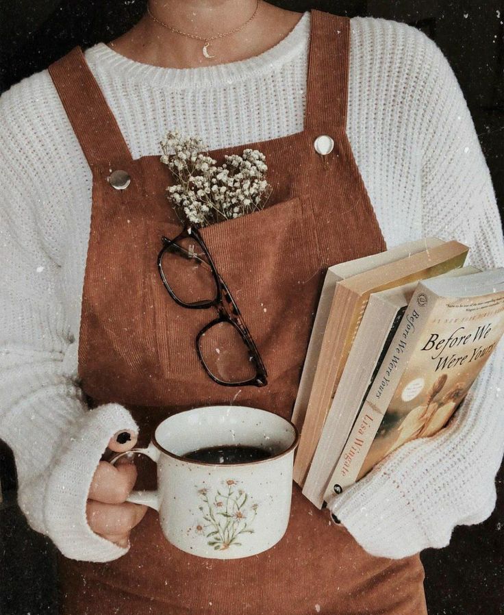 Cozy autumn vibes: Woman holding coffee, books, and glasses in overalls with flowers in pocket. Warm sweater ensemble.