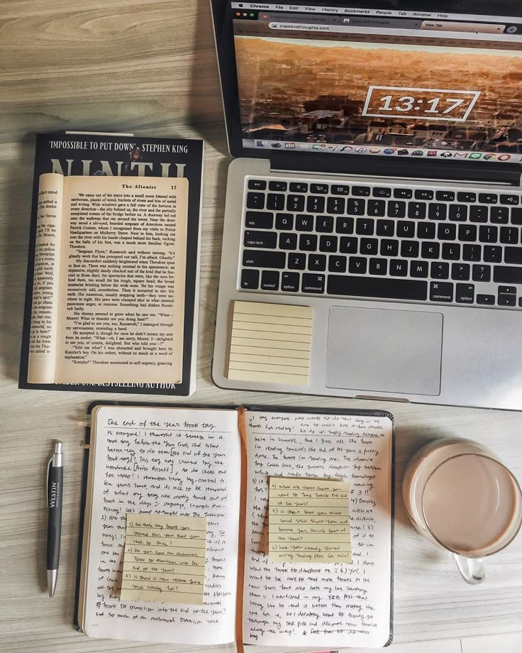 Workspace with open book, laptop displaying time, notebook with notes, pen, sticky notes, and coffee cup on wooden desk.