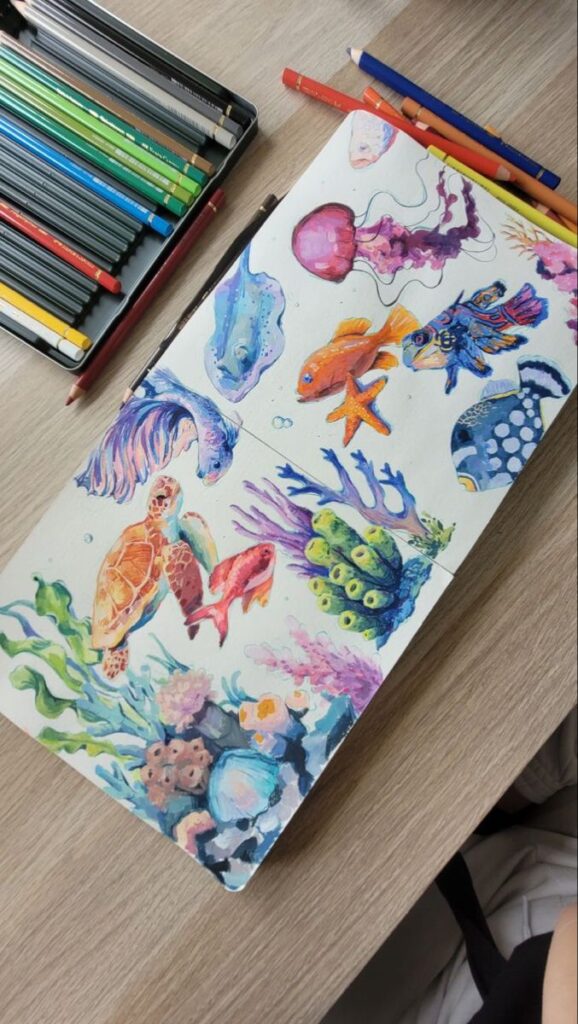 Colorful ocean-themed sketchbook page with sea life illustrations, surrounded by pencils on a wooden table.