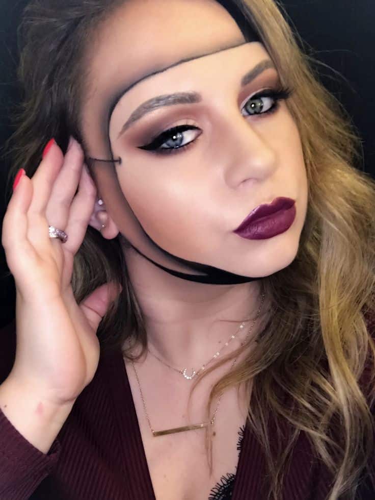 Woman with artistic face paint, bold makeup, and maroon attire, posing with hand near ear.