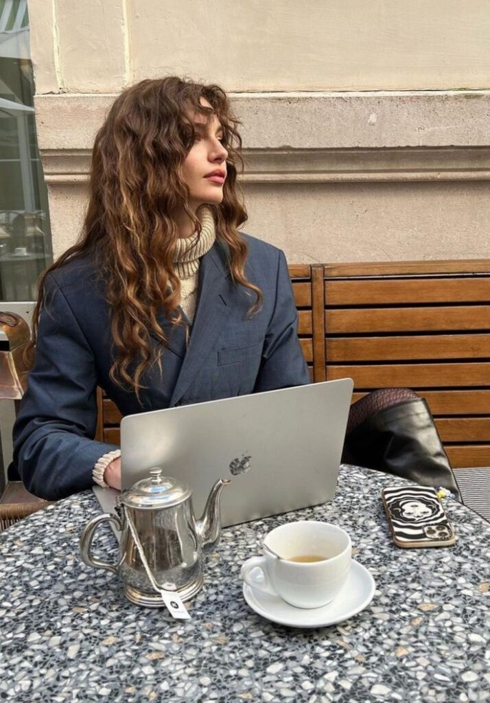 Woman in a blazer sits at a café table with a laptop, tea, and phone, looking away.