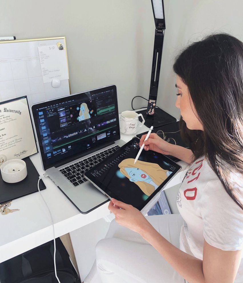 Person designing digital artwork on a tablet next to a laptop in a home office setup.