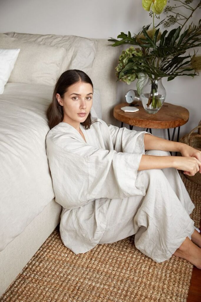Woman in a beige robe sitting on a cozy rug by a sofa, with a wooden table and decorative plants in the background.