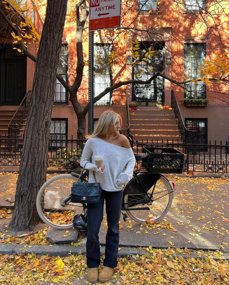 Woman in cozy sweater with coffee by a bicycle, autumn leaves on city sidewalk, warm urban setting.
