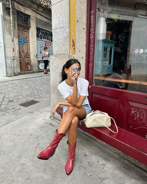 Woman in red cowboy boots sitting outside café, taking a photo with camera, stylish urban street fashion.
