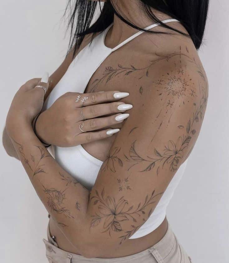 Woman with floral tattoos on arms, wearing a white top and beige pants, showcasing delicate rings and manicure.