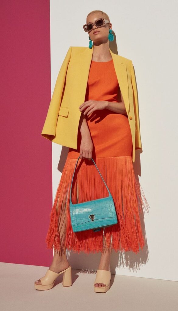 trendy outfit with yellow blazer, orange fringe dress, blue bag, and sunglasses on fashion model standing confidently